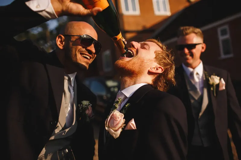 Candid wedding photo of a groom pouring champagne straight from the bottle into the best man's mouth by wedding photographer ARJ Photography®