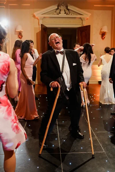 Candid wedding photo of an elderly wedding guest dancing and laughing with two walking sticks by wedding photographer ARJ Photography®