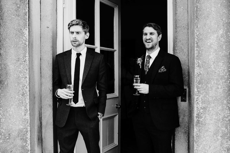 The groom laughs and his friend looks cross while looking at the same thing - powerful black and white wedding photography by ARJ Photography®