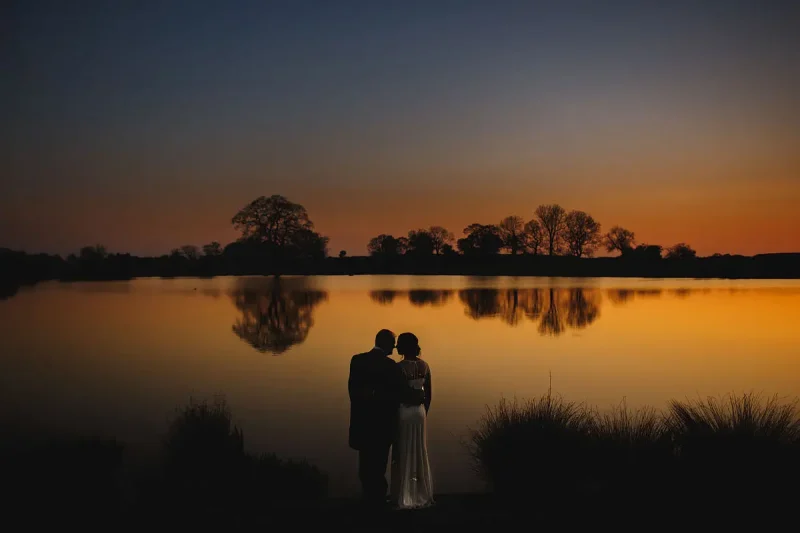 Colourful moody romantic silhouette of a bride and groom by a lake after sunset - artistic wedding photography by ARJ Photography®