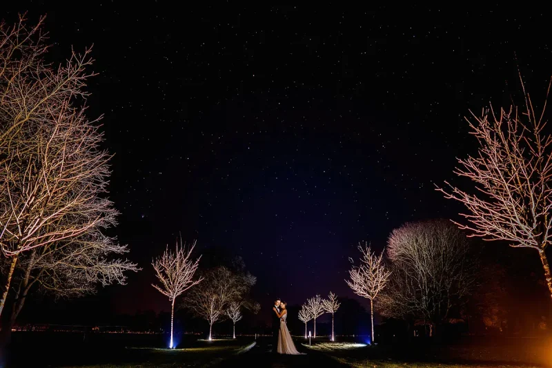 A bride and groom cuddle under a starry night sky surrounded by white trees - artistic wedding photography by ARJ Photography®