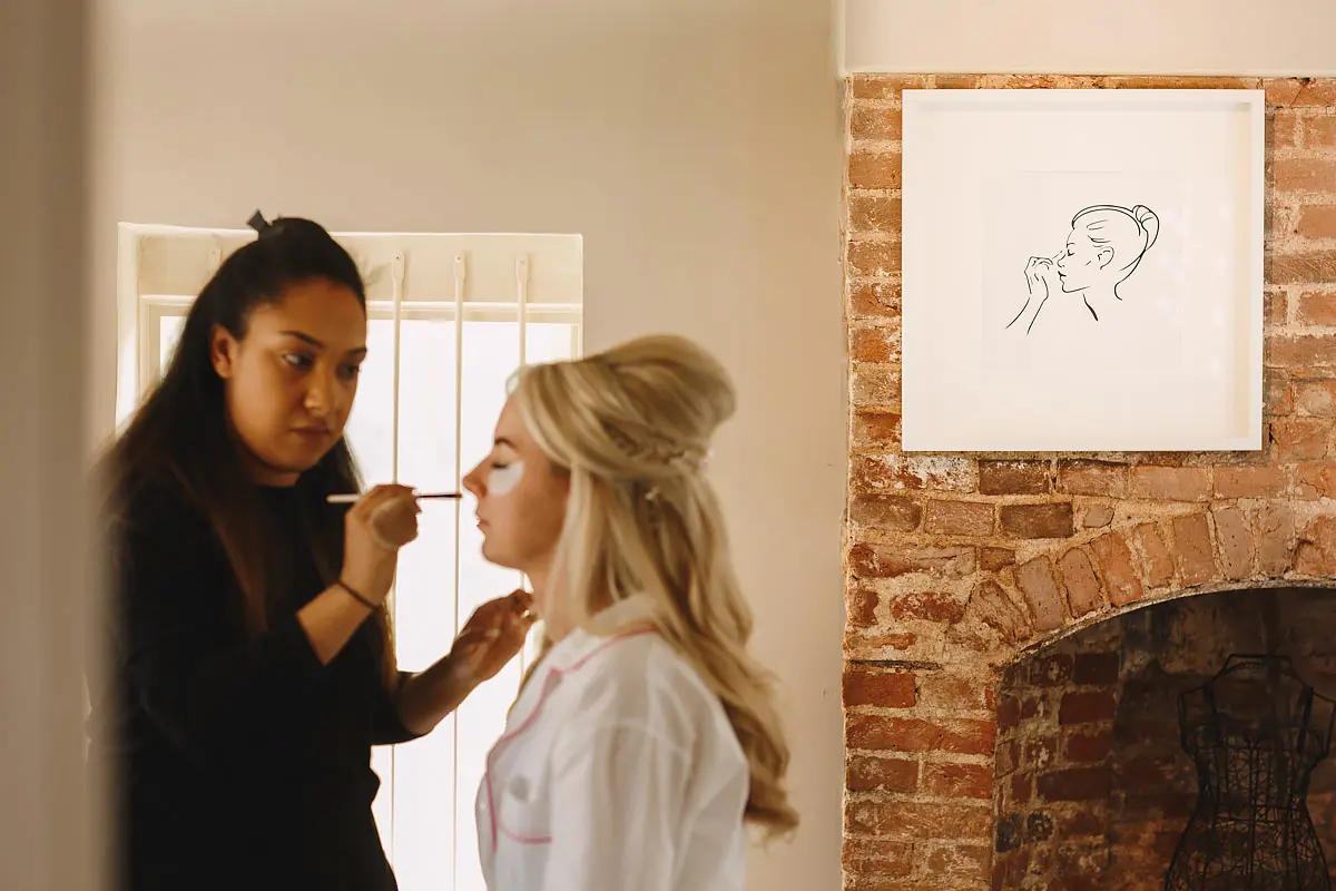 A bride getting ready for her wedding and the makeup artist applying makeup is perfectly mirrored by the picture on the wall - artistic wedding photography by ARJ Photography®