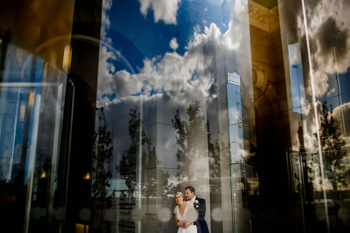Artistic wedding photo of a bride and groom through the window of their wedding venue, with the sky reflected in the window - artistic wedding photography by ARJ Photography®