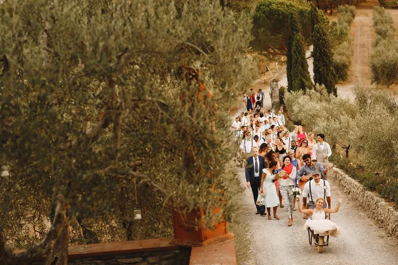 A bride in a wheelbarrow being pushed by the groom and followed by all their guests at a destination wedding in Tuscany Italy by destination wedding photographer ARJ Photography®