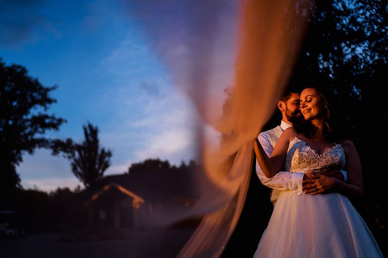Fashion inspired sunset portrait of a bride and groom on their wedding day with the veil blowing in the wind and lit by the setting sun - incredible wedding portraits by ARJ Photography®